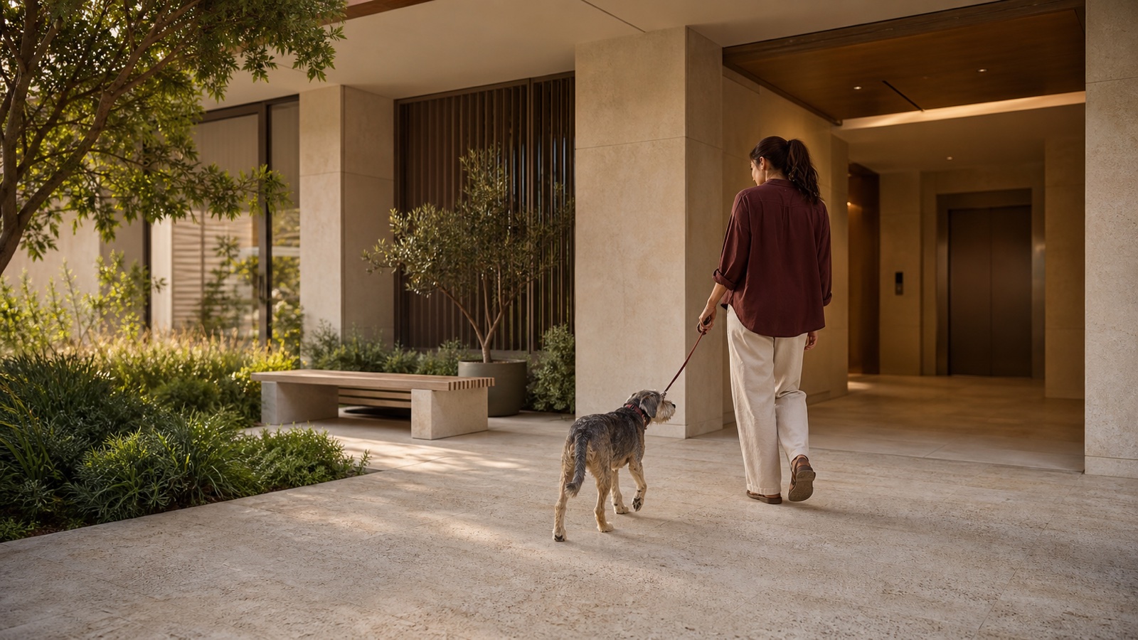 An older small-to-medium dog walks beside a resident on a level shaded route into a calm Dubai apartment building