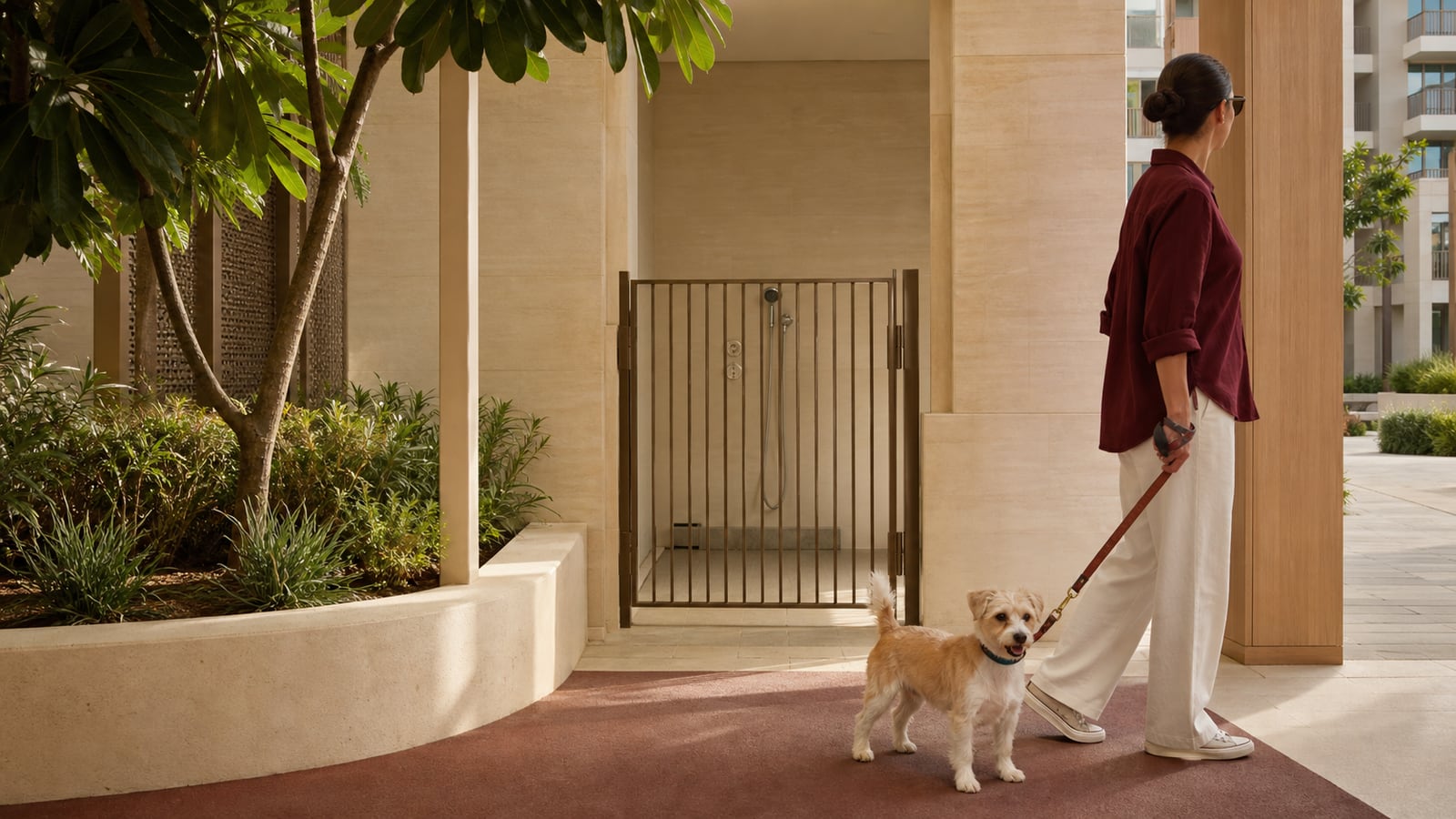 A resident walks a small tan-and-white terrier past a closed gate at a clean shared dog wash point beside a shaded residential courtyard